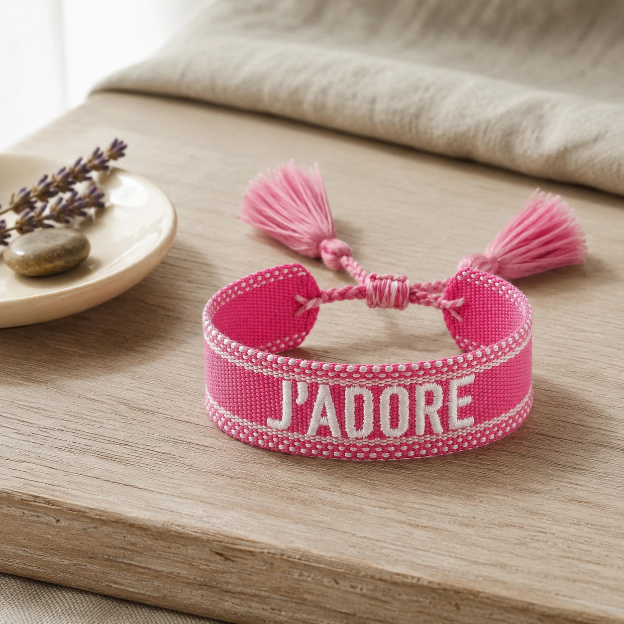 Bracelet tissé de couleur rose avec l'inscription 'J'ADORE', posé sur une planche en bois clair, accompagné d'une petite coupelle contenant des brins de fleurs séchées de lavande et un galet.