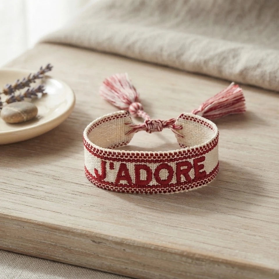 Bracelet tissé de couleur rouge avec l'inscription 'J'ADORE', posé sur une planche en bois clair, accompagné d'une petite coupelle contenant des brins de fleurs séchées de lavande et un galet.