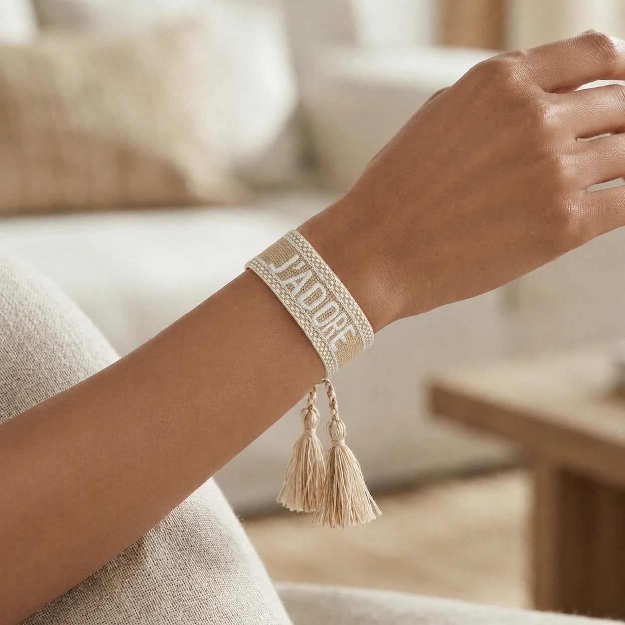Gros plan sur un poignet portant un bracelet tissé de couleur beige avec l'inscription 'J'ADORE' brodée en lettres capitales, agrémenté de deux pompons assortis pendants.