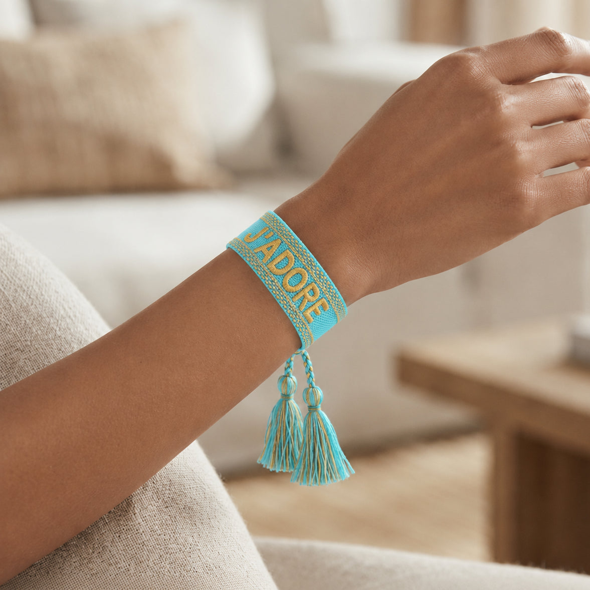 Gros plan sur un poignet portant un bracelet tissé de couleur bleu avec l'inscription 'J'ADORE' brodée en lettres capitales, agrémenté de deux pompons assortis pendants.
