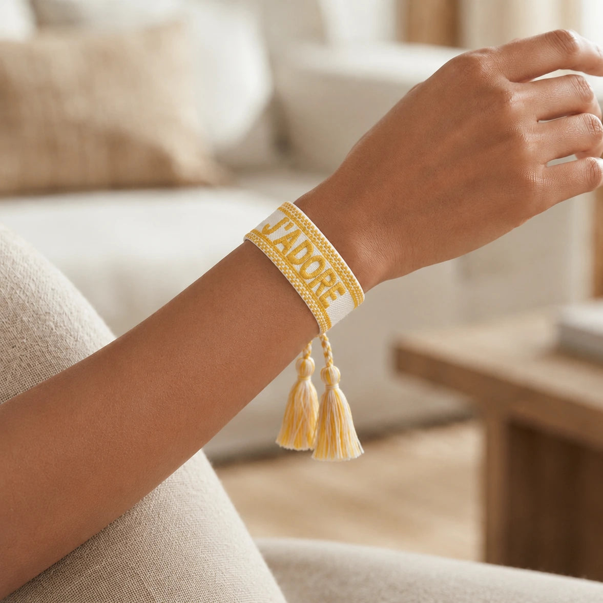 Gros plan sur un poignet portant un bracelet tissé de couleur jaune avec l'inscription 'J'ADORE' brodée en lettres capitales, agrémenté de deux pompons assortis pendants.