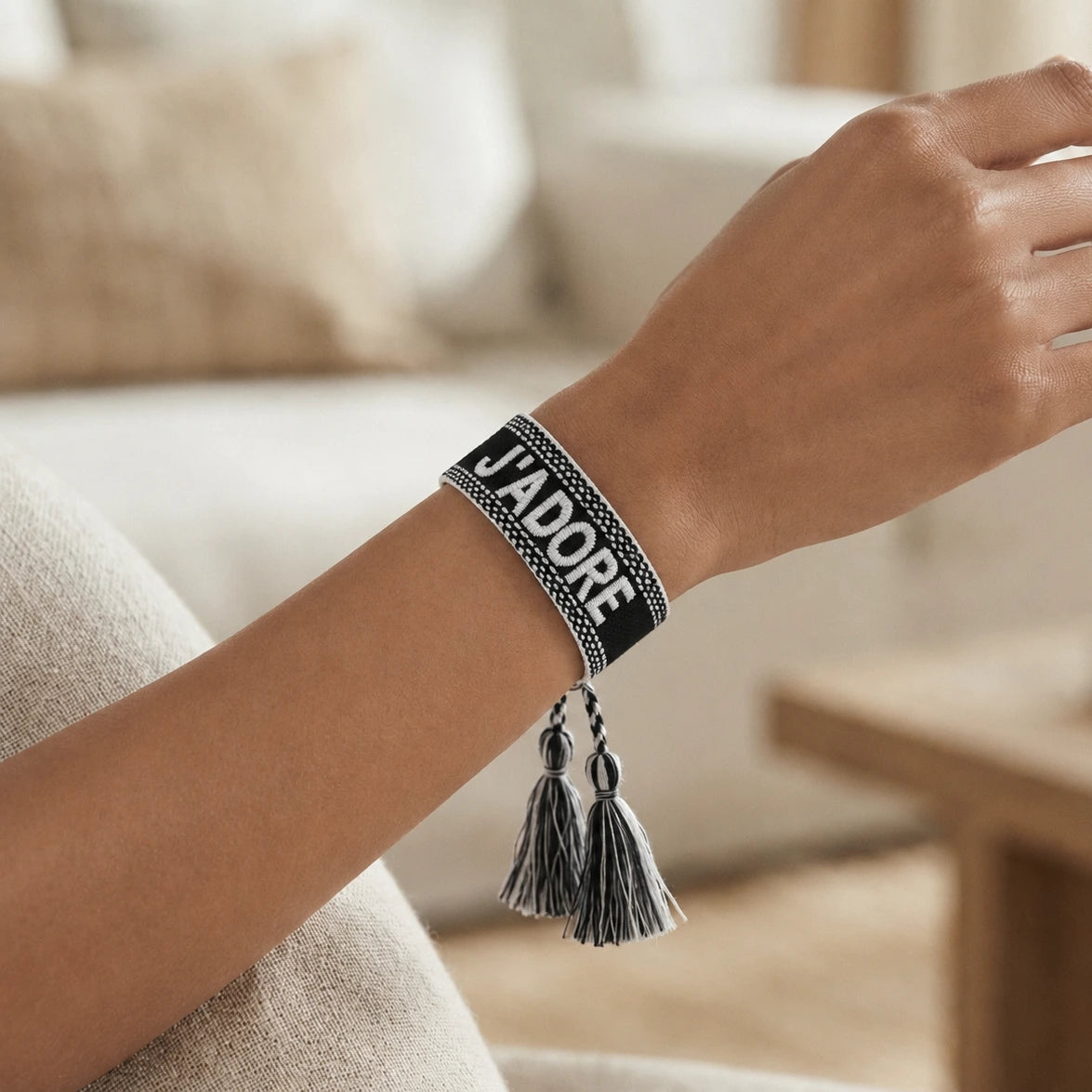 Gros plan sur un poignet portant un bracelet tissé de couleur noir avec l'inscription 'J'ADORE' brodée en lettres capitales, agrémenté de deux pompons assortis pendants.
