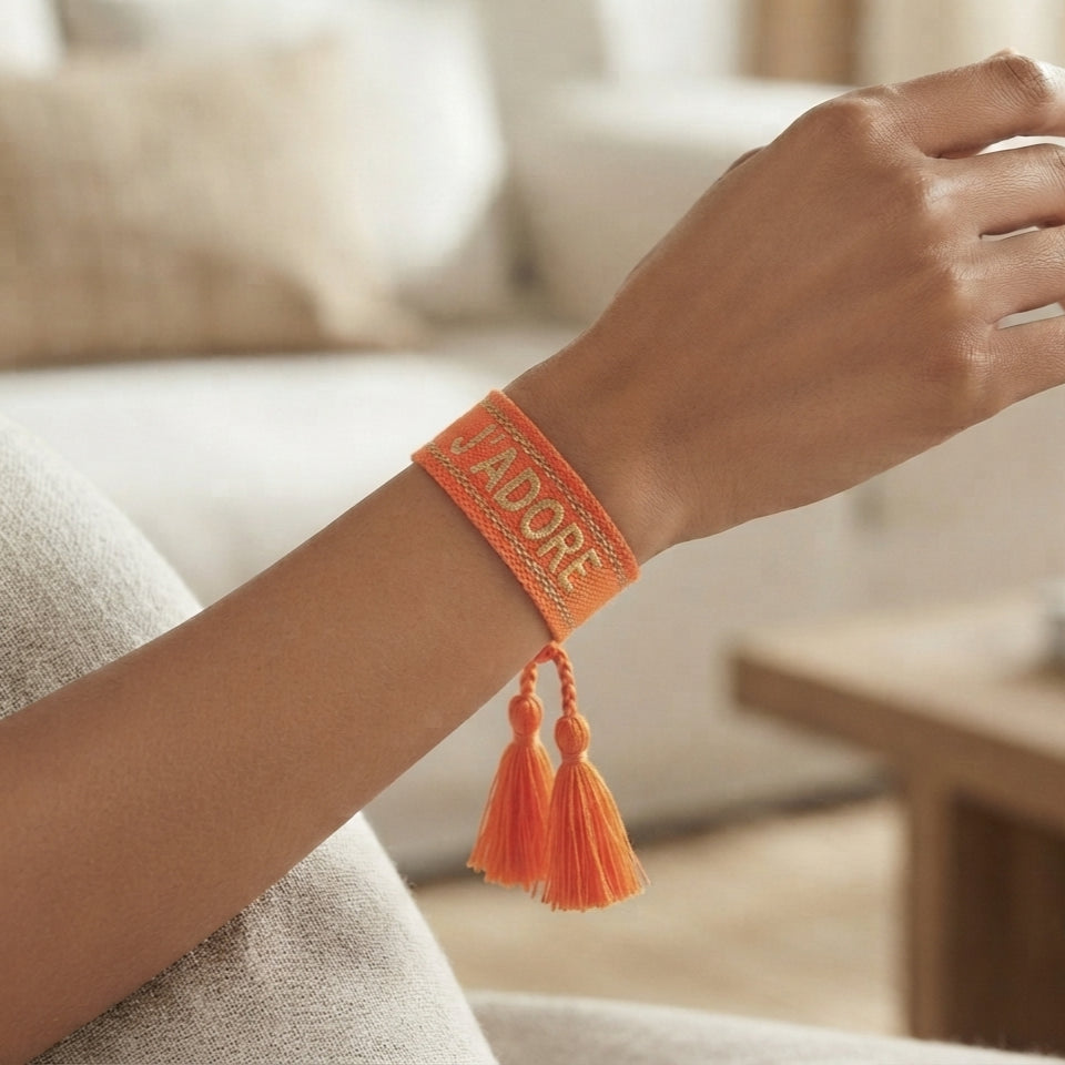 Gros plan sur un poignet portant un bracelet tissé de couleur orange avec l'inscription 'J'ADORE' brodée en lettres capitales, agrémenté de deux pompons assortis pendants.