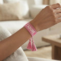 Gros plan sur un poignet portant un bracelet tissé de couleur rose avec l'inscription 'J'ADORE' brodée en lettres capitales, agrémenté de deux pompons assortis pendants.