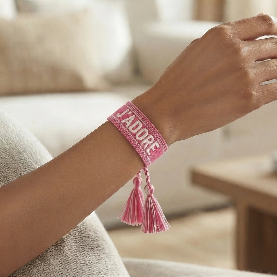 Gros plan sur un poignet portant un bracelet tissé de couleur rose avec l'inscription 'J'ADORE' brodée en lettres capitales, agrémenté de deux pompons assortis pendants.