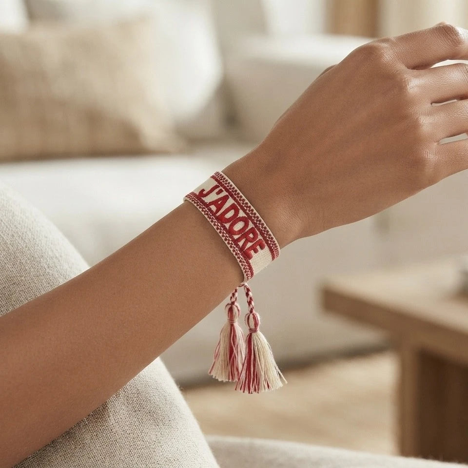 Gros plan sur un poignet portant un bracelet tissé de couleur rouge avec l'inscription 'J'ADORE' brodée en lettres capitales, agrémenté de deux pompons assortis pendants.