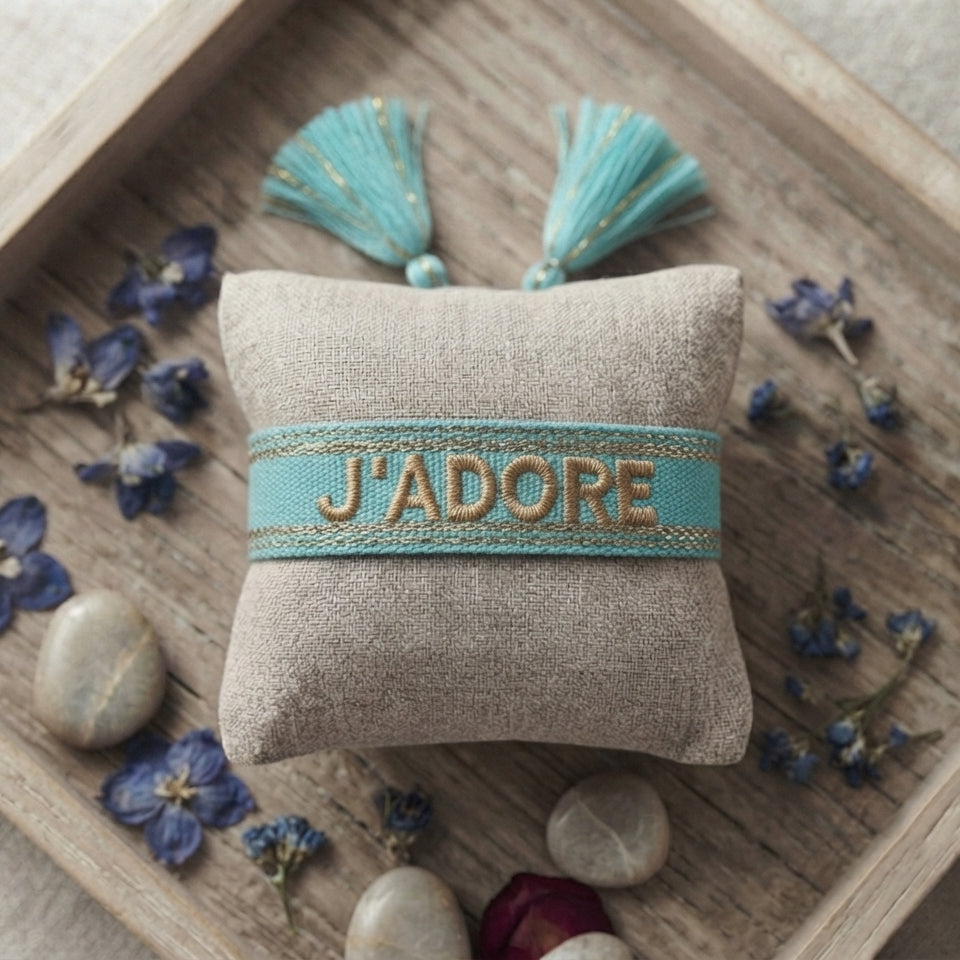 Bracelet tissé de couleur bleu portant l'inscription 'J'ADORE', présenté autour d'un petit coussin en lin beige, disposé sur un plateau en bois rustique avec des pétales de fleurs séchées bleu et des galets décoratifs.
