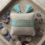 Bracelet tissé de couleur bleu portant l'inscription 'J'ADORE', présenté autour d'un petit coussin en lin beige, disposé sur un plateau en bois rustique avec des pétales de fleurs séchées bleu et des galets décoratifs.