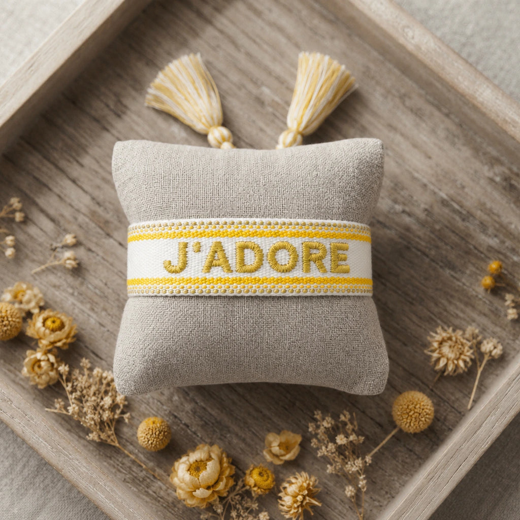 Bracelet tissé de couleur jaune portant l'inscription 'J'ADORE', présenté autour d'un petit coussin en lin beige, disposé sur un plateau en bois rustique avec des pétales de fleurs séchées jaune.