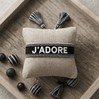 Bracelet tissé de couleur noir portant l'inscription 'J'ADORE', présenté autour d'un petit coussin en lin beige, disposé sur un plateau en bois rustique avec des éléments décoratifs.