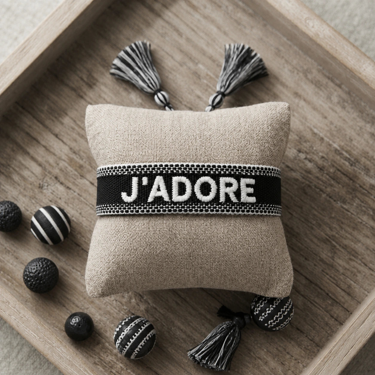 Bracelet tissé de couleur noir portant l'inscription 'J'ADORE', présenté autour d'un petit coussin en lin beige, disposé sur un plateau en bois rustique avec des éléments décoratifs.