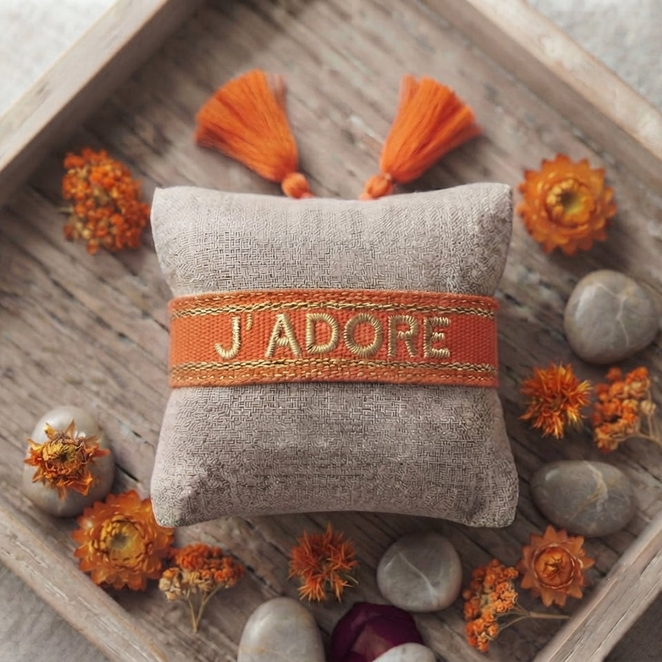 Bracelet tissé de couleur orange portant l'inscription 'J'ADORE', présenté autour d'un petit coussin en lin beige, disposé sur un plateau en bois rustique avec des pétales de fleurs séchées orange et des galets décoratifs.