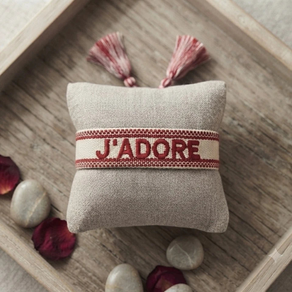 Bracelet tissé de couleur rouge portant l'inscription 'J'ADORE', présenté autour d'un petit coussin en lin beige, disposé sur un plateau en bois rustique avec des pétales de fleurs séchée rouge et des galets décoratifs.