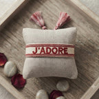 Bracelet tissé de couleur rouge portant l'inscription 'J'ADORE', présenté autour d'un petit coussin en lin beige, disposé sur un plateau en bois rustique avec des pétales de fleurs séchée rouge et des galets décoratifs.