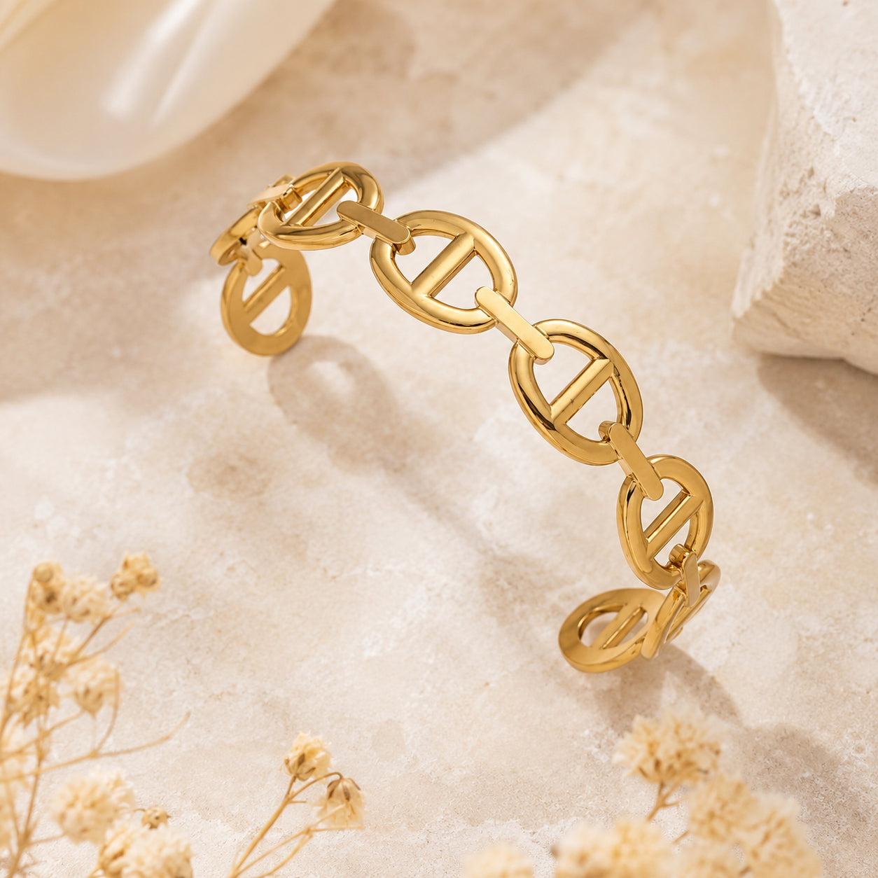 Bracelet jonc Clara doré à maillons style maille marine, présenté sur un support minéral beige avec des fleurs séchées.
