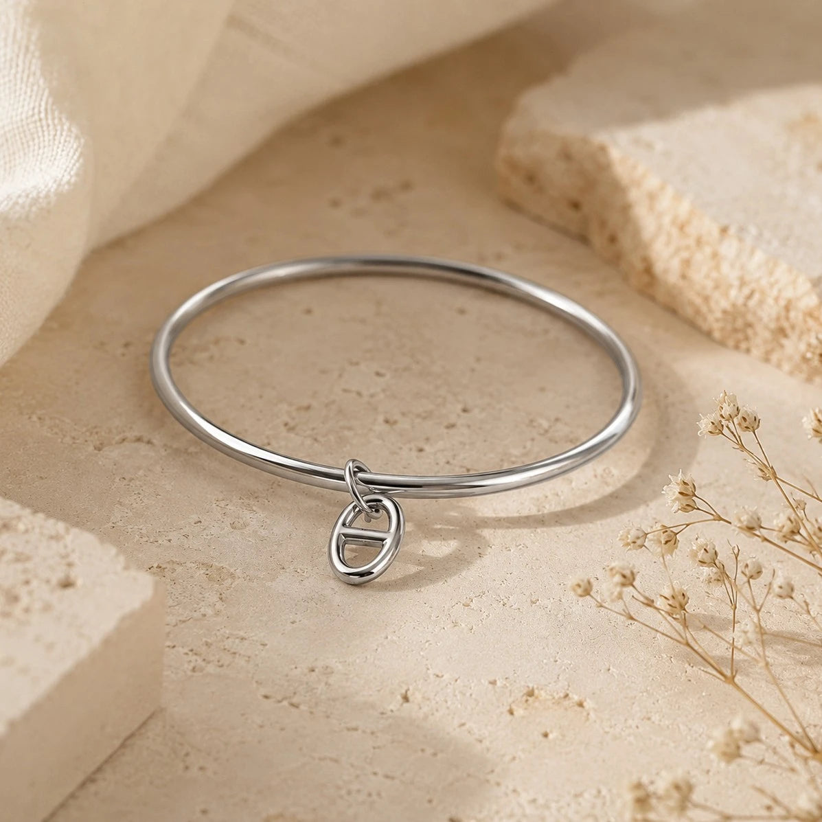 Bracelet jonc Stella en argent avec un pendentif maille marine, posé sur un support en pierre beige entouré de fleurs séchées.