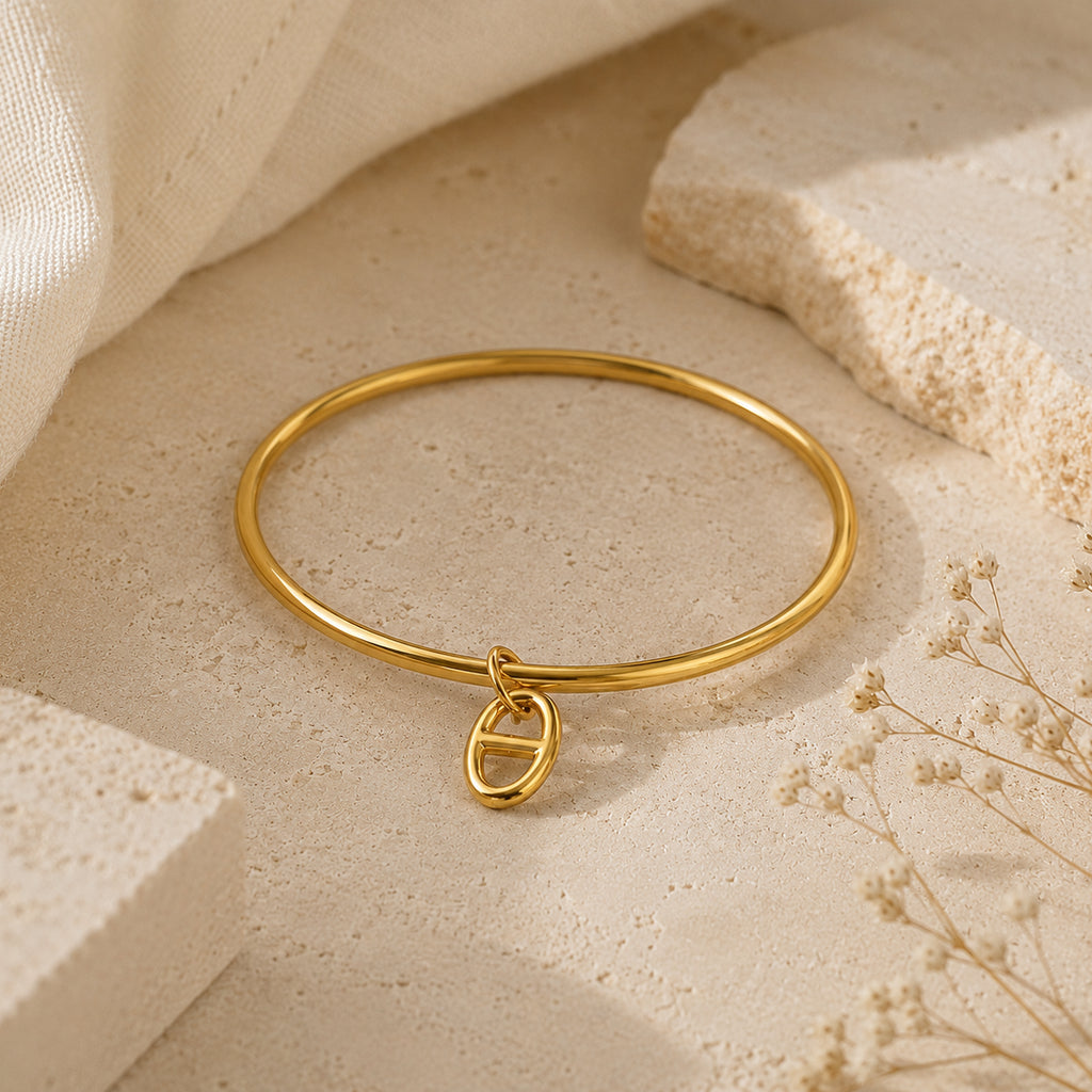 Bracelet jonc Stella doré avec pendentif maille marine, posé sur une pierre travertin beige entourée de fleurs séchées.