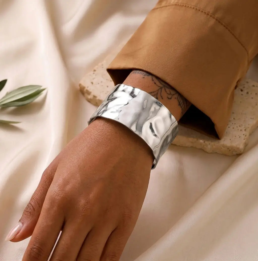 Un bracelet manchette argenté martelé est porté au poignet d'un mannequin à la peau sombre. Le mannequin porte une chemise marron et l'arrière-plan est un tissu de lin texturé.