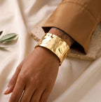 Gros plan d'un bracelet manchette doré martelé, porté au poignet d'un mannequin à la peau sombre. Le mannequin porte une chemise marron et l'arrière-plan est un tissu de lin texturé.