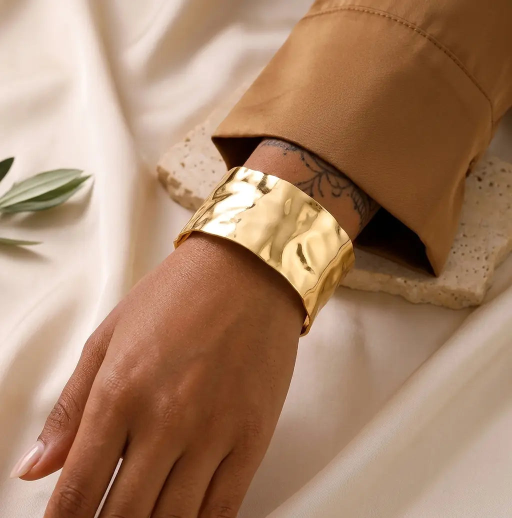 Gros plan d'un bracelet manchette doré martelé, porté au poignet d'un mannequin à la peau sombre. Le mannequin porte une chemise marron et l'arrière-plan est un tissu de lin texturé.