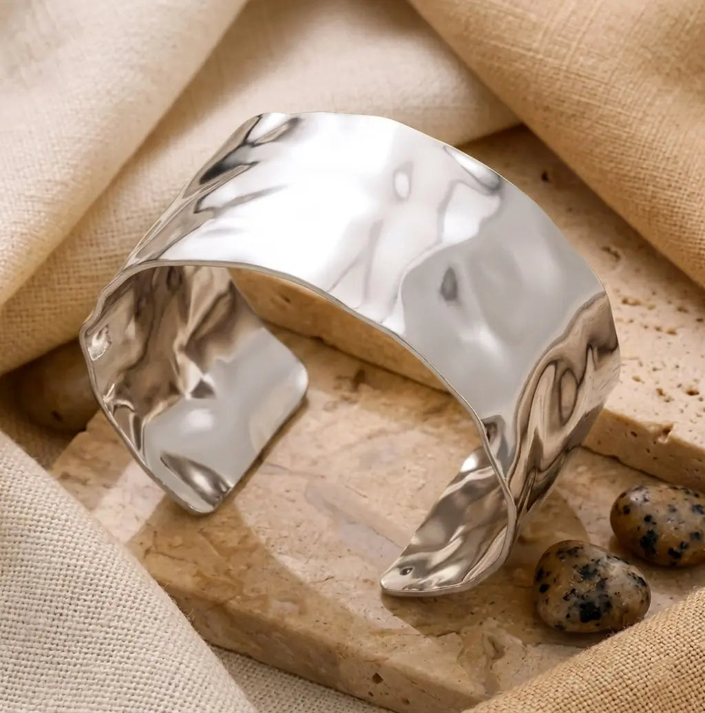 Gros plan d'un bracelet manchette argenté martelé posé sur des dalles de pierre travertine. L'arrière-plan est un tissu de lin texturé et deux petites pierres décoratives tachetées sont visibles à côté.