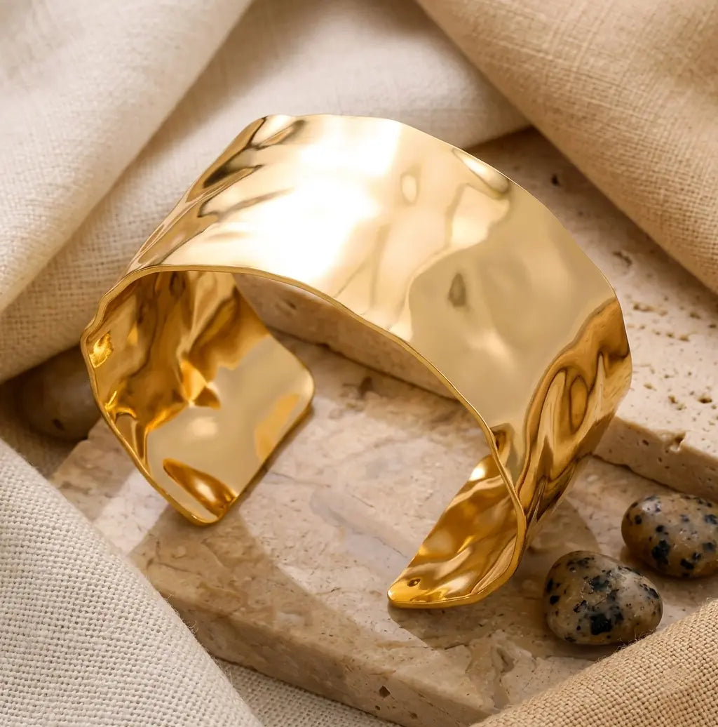 Gros plan d'un bracelet manchette doré martelé posé sur des dalles de pierre travertine. L'arrière-plan est un tissu de lin texturé et deux petites pierres décoratives tachetées sont visibles à côté.