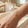 Bracelet Sia - Cristalline en finition or porté au poignet. La fine chaîne dorée et le trèfle scintillant contrastent élégamment sur la peau, avec un arrière-plan flou suggérant un intérieur raffiné.