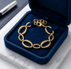 Mise en scène luxueuse du bracelet doré Victoire Vibrante, présenté dans un écrin de velours bleu nuit ouvert. L'écrin repose sur du marbre blanc. Un stylo plume argenté est visible en arrière-plan.