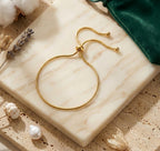 Composition élégante du bracelet de cheville Sienne Infinie doré sur un bloc de marbre, entouré de lavande et d'un pochon vert émeraude.