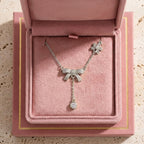 Le collier Adeline la Charmante en argent fin, orné de motifs nœuds et d'une pampille brillante, présenté dans son écrin rose poudré ouvert. 