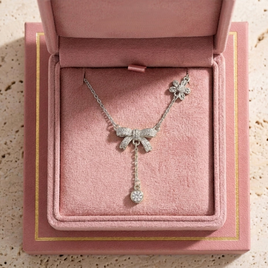 Le collier Adeline la Charmante en argent fin, orné de motifs nœuds et d'une pampille brillante, présenté dans son écrin rose poudré ouvert. 
