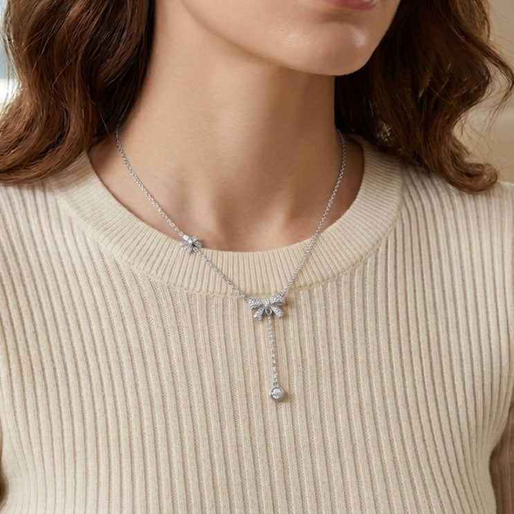 Gros plan d'une femme portant le collier Adeline la Charmante en version argenté, orné de motifs nœuds et d'une pampille brillante, sur un pull côtelé crème.