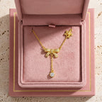 Le collier Adeline la Charmante en or fin, orné de motifs nœuds et d'une pampille brillante, présenté dans son écrin rose poudré ouvert.