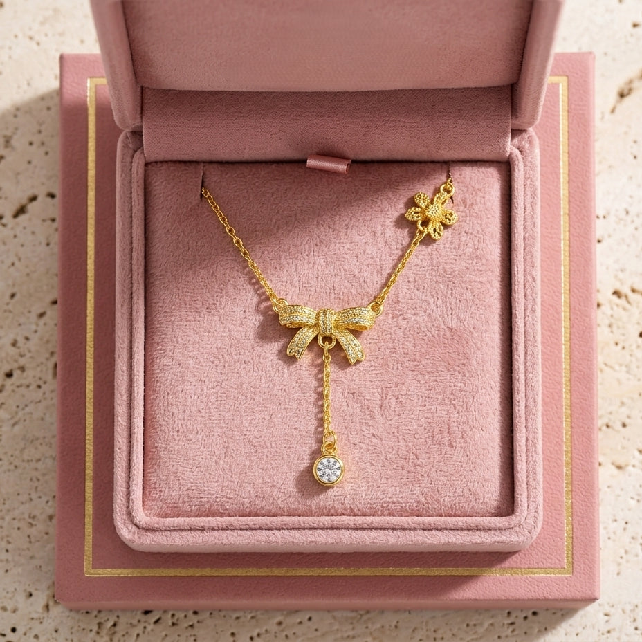 Le collier Adeline la Charmante en or fin, orné de motifs nœuds et d'une pampille brillante, présenté dans son écrin rose poudré ouvert.