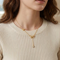 Gros plan d'une femme portant le collier Adeline la Charmante en version dorée, orné de motifs nœuds et d'une pampille brillante, sur un pull côtelé crème.