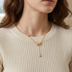 Gros plan d'une femme portant le collier Adeline la Charmante en version dorée, orné de motifs nœuds et d'une pampille brillante, sur un pull côtelé crème.