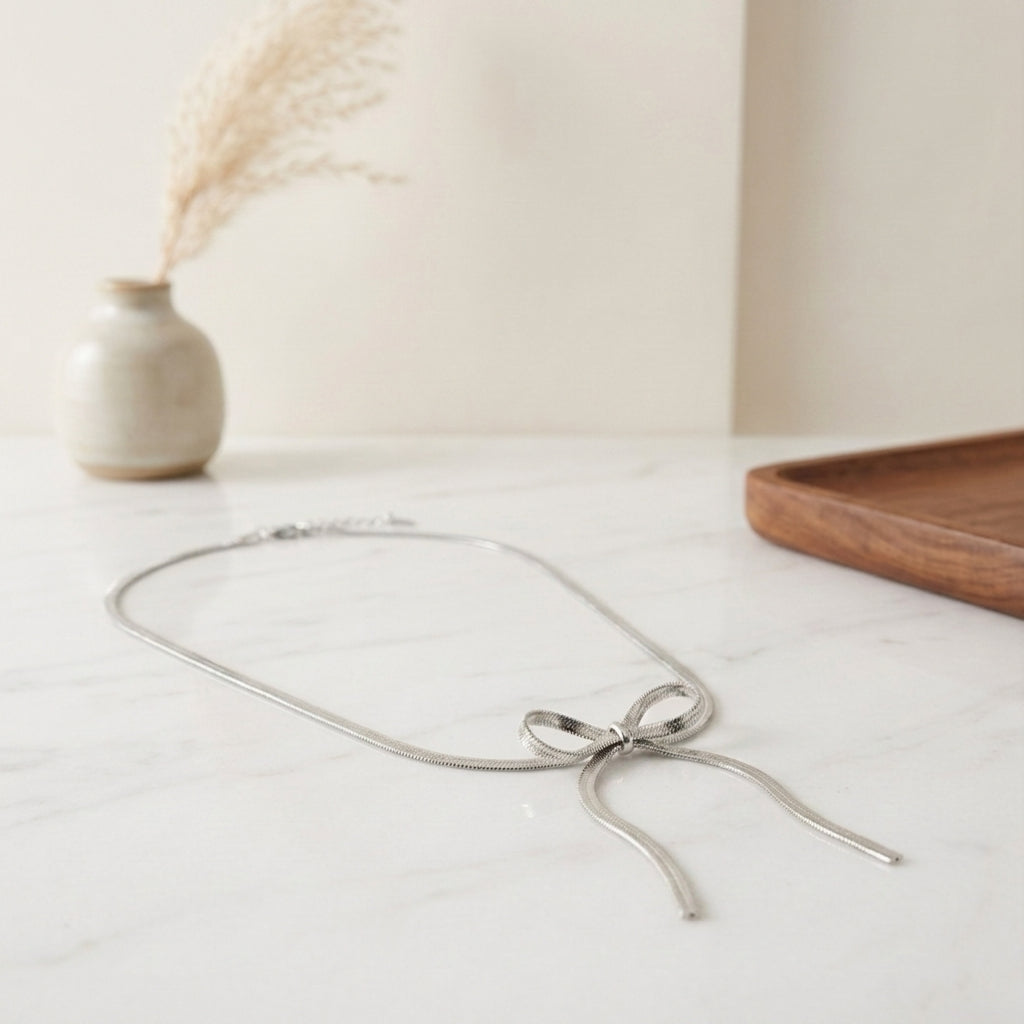 Collier en maille serpent plate argenté formant un nœud délicat, disposé sur une table en marbre blanc dans un décor épuré.
