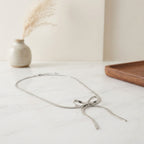Collier en maille serpent plate argenté formant un nœud délicat, disposé sur une table en marbre blanc dans un décor épuré.