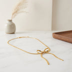 Collier en maille serpent plate dorée formant un nœud délicat, disposé sur une table en marbre blanc dans un décor épuré.