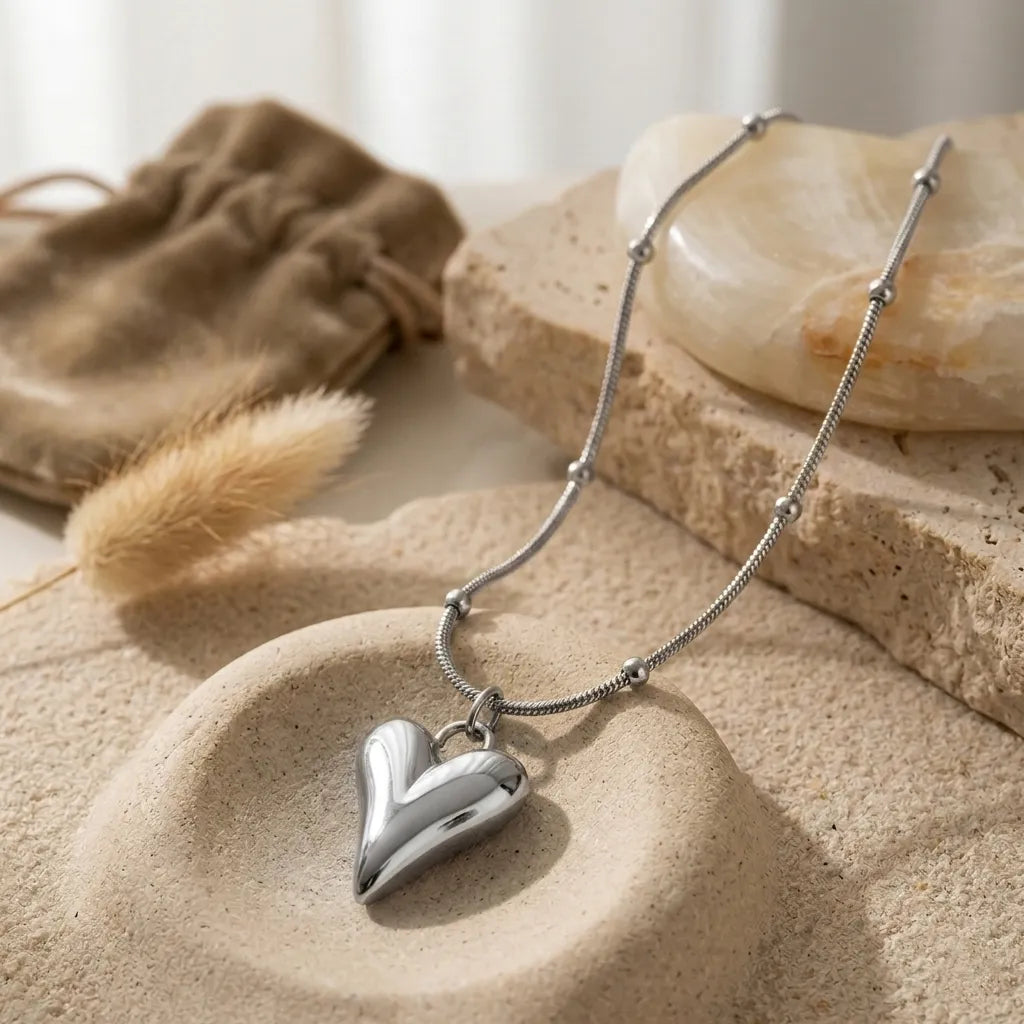 Nature morte du collier Yuna Romantique en argent posé sur des pierres naturelles et du sable fin. Le pendentif cœur volumineux capte la lumière naturelle à côté d'un pochon en velours beige et d'une herbe de la pampa.