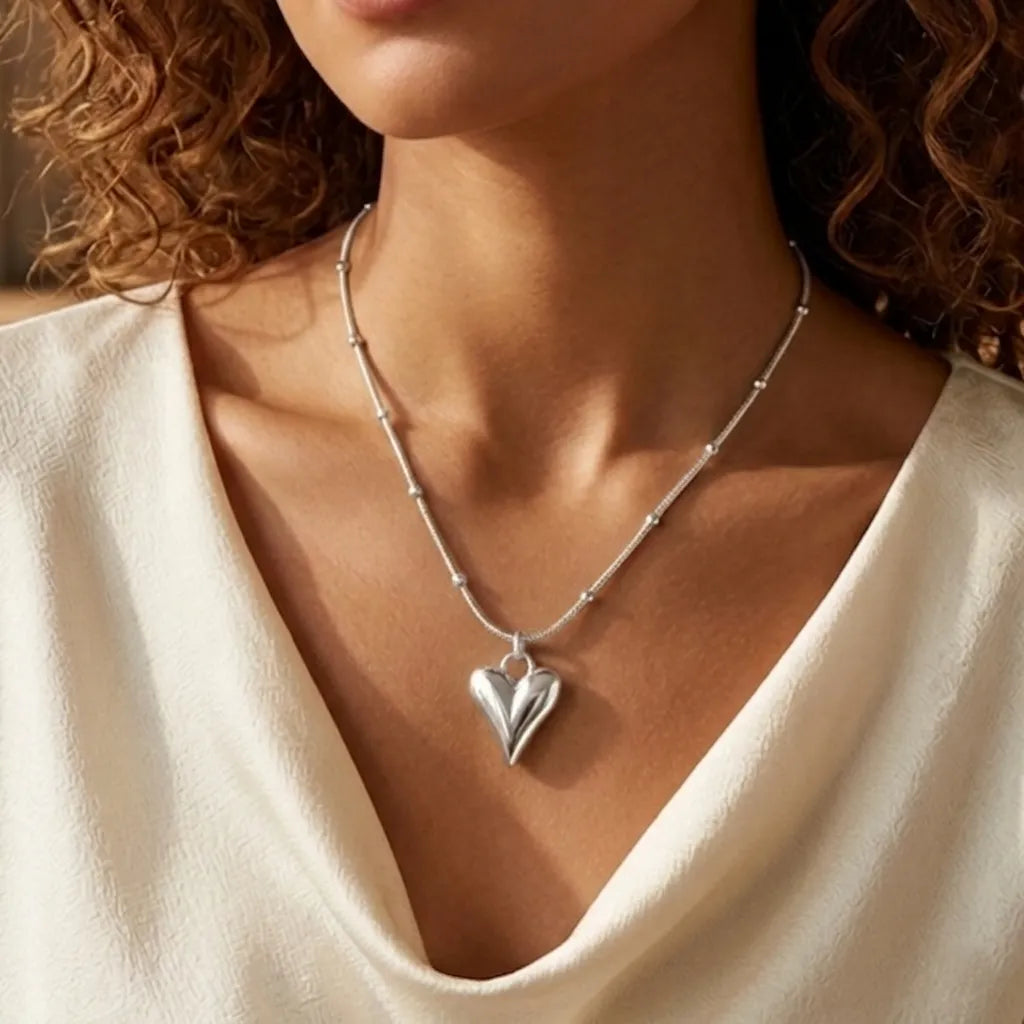 Close-up d'une mannequin portant le collier Yuna Romantique en argent. Le pendentif en forme de cœur bombé et poli reflète la lumière, suspendu à une fine chaîne perlée dorée sur un haut drapé blanc élégant.