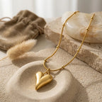 Nature morte du collier Yuna Romantique en or posé sur des pierres naturelles et du sable fin. Le pendentif cœur volumineux capte la lumière naturelle à côté d'un pochon en velours beige et d'une herbe de la pampa.