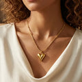 Close-up d'une mannequin portant le collier Yuna Romantique en or. Le pendentif en forme de cœur bombé et poli reflète la lumière, suspendu à une fine chaîne perlée dorée sur un haut drapé blanc élégant.
