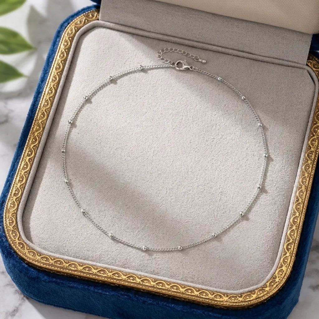 Image du collier Yzée argenté soigneusement présenté dans un écrin de luxe ouvert. Le bijou en acier argenté est disposé en boucle sur le fond en velours gris perle de la boîte, encadré par le bord en velours bleu roi et la frise dorée de l'écrin. Une présentation parfaite pour un cadeau précieux et intemporel.