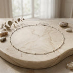 Photographie de style nature morte du collier Yzée en version argenté, posé en cercle sur un plateau en marbre beige veiné. Le bijou, une fine chaîne à boules, est entouré de galets naturels lisses et d'une branche d'eucalyptus séchée. La lumière douce vient d'une fenêtre floue en arrière-plan, créant une ambiance élégante et sereine.