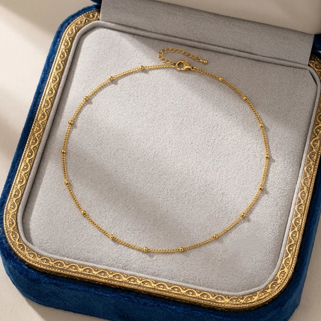 Image du collier Yzée doré soigneusement présenté dans un écrin de luxe ouvert. Le bijou en acier argenté est disposé en boucle sur le fond en velours gris perle de la boîte, encadré par le bord en velours bleu roi et la frise dorée de l'écrin. Une présentation parfaite pour un cadeau précieux et intemporel.