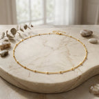 Photographie de style nature morte du collier Yzée en version dorée, posé en cercle sur un plateau en marbre beige veiné. Le bijou, une fine chaîne à boules, est entouré de galets naturels lisses et d'une branche d'eucalyptus séchée. La lumière douce vient d'une fenêtre floue en arrière-plan, créant une ambiance élégante et sereine.