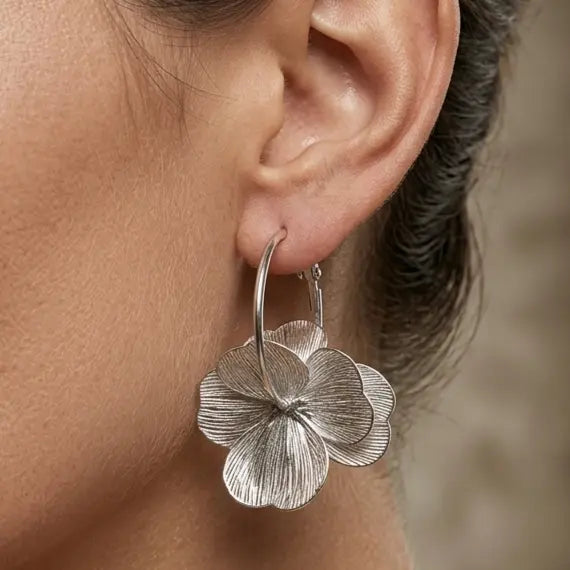gros plan sur une grande créole pendante Eléonore de Maison Dosana, en acier inoxydable argenté brossé, ornée d'une fleur texturée généreuse, portée à l'oreille.