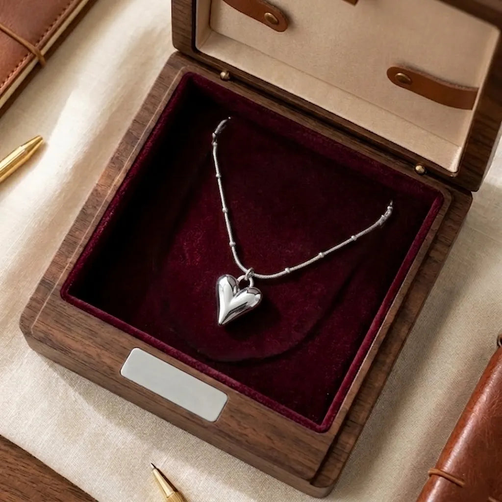Collier Yuna Romantique argenté présenté dans un luxueux écrin en bois foncé avec un intérieur en velours bordeaux. La chaîne est élégamment disposée pour montrer le pendentif cœur poli.