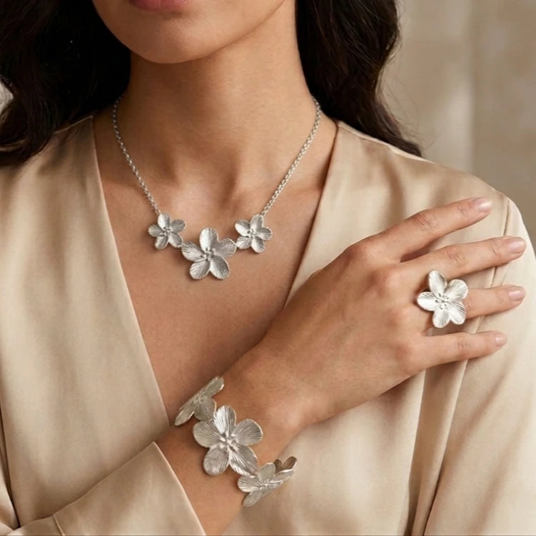 Portrait rapproché d'une femme portant la parure complète en argent Colette - Épanouie. Le collier à trois fleurs, la bague imposante et le bracelet manchette assorti présentent des pétales finement ciselés qui capturent la lumière sur une tenue en satin.